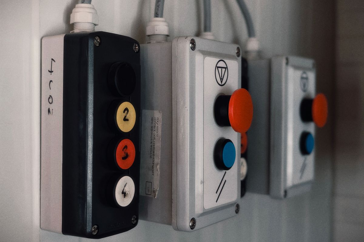 Trois boîtiers de commande industriels alignés sur un mur, boutons-poussoirs rouges, bleus et jaunes.