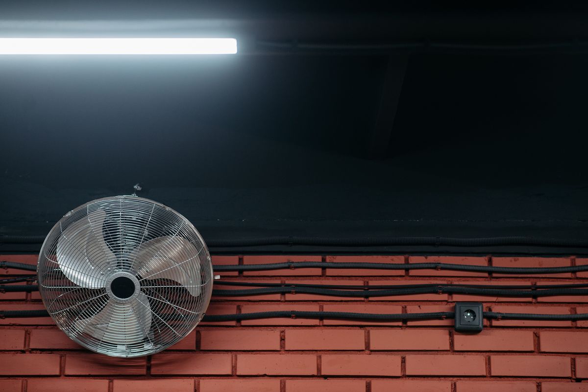 Ventilateur industriel métallique fixé sur un mur de briques, sous un tube fluorescent, gaines électriques apparentes.